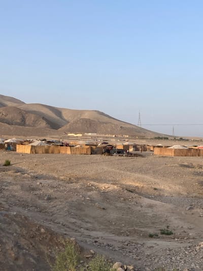 A wide-angle image captures an informal settlement within an arid, rocky desert landscape in Spezand, Pakistan. The settlement, located in the mid-ground, consists of numerous makeshift dwellings constructed from light-colored woven materials or reeds for walls and white or light-colored tarps for roofs, arranged in clusters. A red tractor is visible within the settlement, positioned towards the right of a central group of structures, with several small, indistinct human figures present nearby. Piles of refuse or organic material are observed around some dwellings. The foreground consists of barren, uneven, rocky, and dusty ground with sparse, low-lying scrub vegetation. In the background, light-brown, undulating barren hills or low mountains dominate, under a clear, pale blue sky. Two metallic lattice power transmission towers are visible on the distant hill slopes on the right, with power lines stretching horizontally across the scene. Lighting indicates daylight conditions.