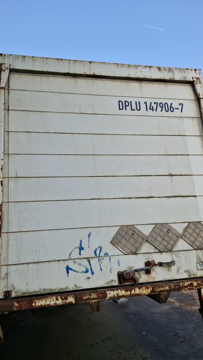 An exterior low-angle shot captures a weathered, white shipping container against a clear blue sky. The container's surface is marked by horizontal lines and stained with dirt and rust, particularly along the bottom edge and frame. A shipping identification number, "DPLU 147906-7," is stenciled in dark blue on the upper right portion of the container. Below this, blue graffiti is visible, along with three diamond-shaped metal plates. A rusty latch mechanism is attached to the lower right side of the container. The foreground shows a dark, wet surface, possibly asphalt, with reflections. A thin contrail is visible in the sky. The image was taken in Ichenhausen, Germany, near the railway tracks.