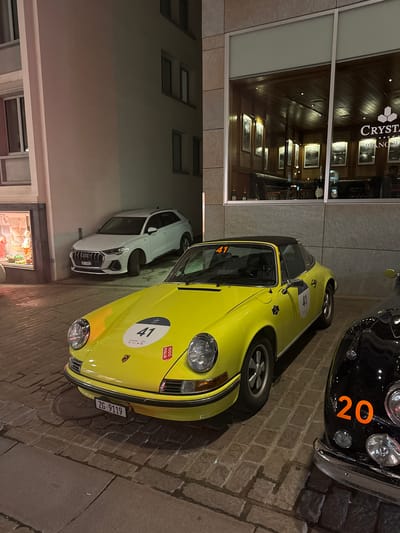 The scene depicts an outdoor urban environment at night in Sankt Moritz, Switzerland, with several vehicles parked on a cobblestone surface. In the foreground, a bright yellow classic Porsche 911 Targa is prominently featured, facing forward. It displays rally number "41" on its hood, windshield, and driver's side door, alongside "Chopard" and "1000 MIGLIA" event decals. The vehicle's Swiss license plate is "ZG-9119".

To the immediate right of the Porsche, the front portion of a black classic car with an orange rally number "20" on its fender is partially visible. Further in the background, to the left of the yellow Porsche, a white Audi Q3 SUV with Swiss license plate "GR-51014" is parked within a building alcove next to a ground-level retail display window containing mannequins.

The building on the right features a large window revealing the interior of the "CRYSTAL PIANO BAR," identified by visible signage. This interior is richly appointed with dark wood paneling on walls and ceiling, recessed lighting, framed artworks, and dark leather seating. Bottles, possibly champagne, are visible on a surface near the window, and a faint human silhouette can be discerned deeper within the bar. The cobblestone pavement exhibits wet reflections in several areas.