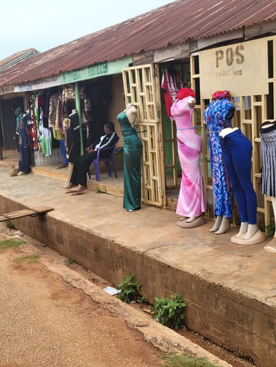 A street-level view of a market stall in Tangu, Nigeria. The stall displays an assortment of clothing on mannequins and shelves. In the foreground, a series of mannequins are dressed in brightly colored garments, including a pink, figure-hugging dress, a blue patterned dress, and a blue pair of leggings. Behind the mannequins, shelves are stacked with fabric and folded clothing. To the left, the corrugated metal roof of another stall is visible. A sign above the displayed clothing reads "POS AVAILABLE HERE". Further back, a person is seen working at the stall. The ground in front of the stall is a concrete pathway. The scene appears to be outdoors, with natural light suggesting daytime. A small patch of green plants grows alongside a drainage ditch to the right of the stall. The overall impression is one of a busy commercial area.