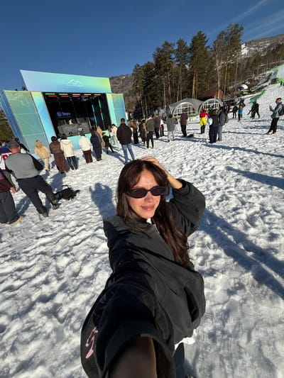 A woman with long dark hair and sunglasses lies on snow, taking a selfie with a crowd of people and a stage in the background. The stage features a large screen and the words "Malinovka Fest" in Russian. The location appears to be Озёрное, Russia, as the festival is known to take place there. The sky is clear and blue.
