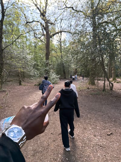 The image is a horizontally oriented photograph, showing an outdoor scene rotated 90 degrees clockwise. In the bottom-right corner, the right hand and forearm of a person with dark skin are visible. The hand is making a peace sign and holds a clear plastic water bottle. A silver-toned wristwatch with a distinct rectangular case, a round light blue or grey dial featuring two sub-dials, and a metallic strap is worn on the wrist. The label on the water bottle is partially visible, showing "0.5 Litr[e]" and the brand "Minar Water".

The main portion of the image displays a dirt and gravel path winding diagonally from the mid-right towards the top-left. Several individuals are walking along this path, moving away from the camera. The person closest to the foreground is wearing a black hoodie, black trousers, and white sneakers. Further ahead, another person is dressed in a white top and dark bottoms. More blurry figures are discernible in the distance, with one person wearing a blue patterned jacket.

The path is bordered by numerous trees, many of which are adorned with new, light green foliage, suggesting a spring or early summer season. The sky, appearing bright and somewhat hazy, can be seen through the tree canopy on the left side of the image, while dense tree cover occupies the upper sections. The lighting indicates it is daytime. The scene depicts a leisurely walk or stroll through a natural, wooded environment or park, consistent with settings found in Poole, United Kingdom. The peace sign gesture by the foreground subject adds a relaxed and positive element to the photograph.