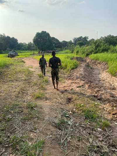 The image displays a rural scene in Ayati, Nigeria, under a bright, partially cloudy sky. In the foreground, a dirt path, sparsely covered with dry vegetation and scattered debris, extends into the mid-ground. Two individuals in military-style camouflage uniforms are walking away from the camera along this path. The person closest to the camera is barefoot, wearing a camouflage t-shirt and trousers. The individual slightly ahead and to their left wears a long-sleeved camouflage top, trousers, and appears to be wearing boots.
The path is flanked by lush green fields of cultivated crops on both sides. A distinct excavated trench, showing recent earthworks, runs parallel to the path on the right side.
In the mid-ground, to the far left, a camouflaged military utility vehicle, possibly an armored SUV or pickup, is partially visible within the green fields, positioned off the main path. A mounted weapon system, resembling a heavy machine gun, is visible on top of this vehicle, and several individuals are present on or near it. Further down the path, beyond the walking military personnel, a group of approximately 15-20 civilians, including visible children, is gathered near the edge of a village or settlement, characterized by visible structures and dense trees.
The background features a line of dense green trees, with a large, prominent tree centrally located. A cellular communication tower is partially visible above the treeline on the far right horizon. The lighting suggests late afternoon conditions.