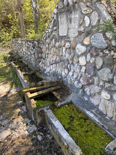 The image depicts a stone structure likely related to a water system, possibly a series of connected reservoirs or channels. The structure is constructed from irregularly shaped stones and concrete. Some sections of the channels are filled with green algae-covered water. Wooden beams are present, possibly for support or division. A stone plaque with indistinct markings is integrated into the stone wall. Vegetation is visible at the periphery of the structure. The location is Boboshevo, Bulgaria.
