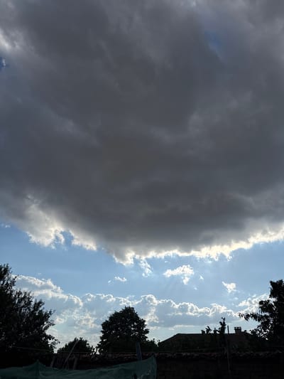 The image shows a dramatic sky scene with dark, heavy clouds dominating the upper portion, casting a shadow over the landscape below. Sunlight breaks through in some areas, creating beams of light that illuminate the lower, brighter clouds. Silhouetted against the sky are the tops of trees and the roofs of a few buildings, suggesting a rural or semi-rural setting. The architecture of the buildings, particularly the tiled roofs and chimneys, is indicative of Bulgarian construction. The scene is devoid of any people or explicit actions. The overall impression is one of approaching or receding weather, with the interplay of light and shadow creating a sense of depth and atmosphere. The location is identified as Blatets, Bulgaria.