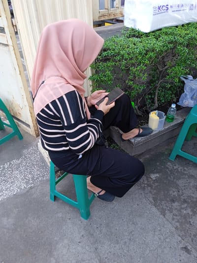A person wearing a salmon-colored hijab, a black and white striped sweater, and black pants is sitting on a green plastic stool. They are looking at a mobile phone they are holding in their hands. The location appears to be outdoors, possibly in Pilang, Indonesia, based on the visible company label and other contextual cues. Beside the person is a small green bush, a clear plastic container with a yellow sponge inside, and a bottle of liquid, likely soap. Another green stool is situated to the right of the person. A white object is behind the bush. The ground is concrete.