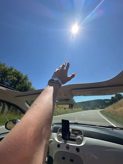 The image depicts a first-person perspective from inside a convertible car, driving on a winding road in Vöhrenbach, Germany. The sun is prominently visible in the clear blue sky, casting bright rays and lens flares. A person's arm is raised, reaching towards the sun, adorned with bracelets and a watch. The car's interior is partially visible, including the dashboard with a phone mount and a sun visor. The surrounding environment shows lush green trees on one side of the road and dry, grassy hills on the other. The road stretches ahead, disappearing into the distance, indicating a scenic drive through the countryside.