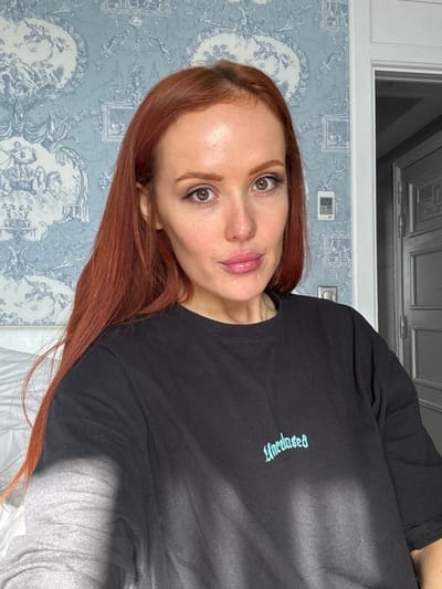 The scene depicts a headshot-style portrait of a single adult female in an indoor setting, located in Deauville, France. The female, centered in the frame, has long, reddish-brown hair, a fair complexion, and is looking directly at the camera. Her lips are full and exhibit a glossy finish. She is wearing a black, long-sleeved t-shirt with the word "Unbased" embroidered in light blue script on the chest. The background consists of a wall covered in blue and white toile de Jouy wallpaper, featuring detailed pastoral or classical vignettes. To the right of the female's head, a white rectangular air conditioning control unit is mounted on the wallpaper. Below it, a light-colored electrical outlet is visible. A white door frame partially visible on the far right leads to an adjacent room, revealing a light-colored, paneled door beyond. White bedding is partially visible in the lower-left corner of the frame.