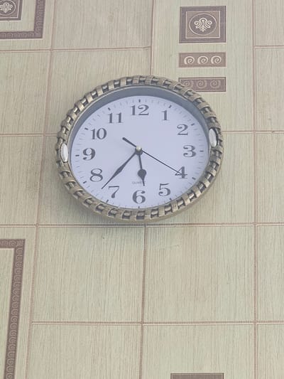 A close-up, straight-on shot shows a round, analog clock with a decorative golden bezel, hanging on a wall. The clock face is white with black numbers and hands. The hour hand is pointing to the 7, and the minute hand is pointing to the 12. The clock is centrally located in the frame. The wall behind the clock has a tiled pattern in shades of beige and brown, with subtle decorative squares and rectangles interspersed. The image appears to be taken indoors, possibly in a residential or public building, with no specific context for its location in Таёжное, Russia provided by the image itself.