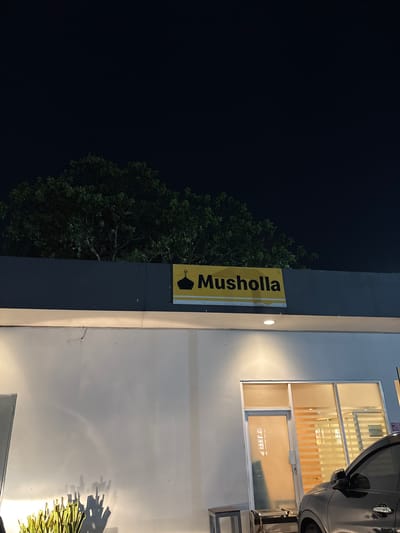 The image shows a vertical yellow sign with the word "Musholla" in black letters, accompanied by a crescent moon and star symbol, affixed to a dark gray wall. Adjacent to the wall, a tree with dense green foliage is visible against a dark night sky. To the right of the gray wall, a white wall with a window reflecting interior lights is present. Part of a gray car is visible parked near the building. The location is Kelapa Dua, Indonesia, as indicated by the signage.
