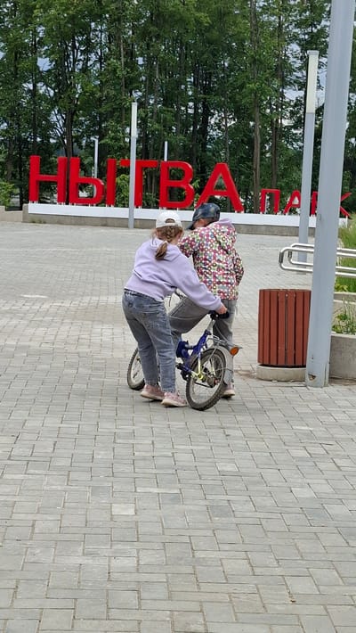 Two children, a girl and a boy, are interacting with a blue bicycle on a paved walkway. The girl, wearing a light purple hooded sweatshirt and blue jeans, is on the left and appears to be helping the boy, who is on the right. The boy is wearing a colorful patterned hooded sweatshirt and a dark baseball cap. He is seated on the bicycle, and the girl has her hands on the handlebars and the back of the bike, suggesting she is either teaching him to ride or assisting him. They are positioned in front of a large red sign that spells out "НЫТВА" (Nytva) in bold, block letters, indicating this is likely a welcome sign or landmark in the city of Nytva, Russia. The background consists of a line of trees and a cloudy sky. To the right of the children, there is a wooden trash receptacle and a silver railing. The ground is paved with light gray rectangular bricks arranged in a repeating pattern.