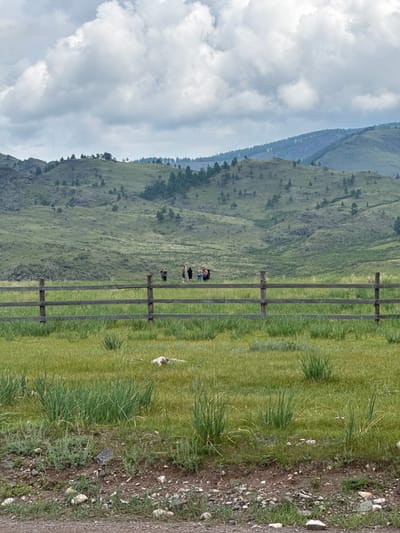 The image depicts a rural landscape with rolling green hills under a cloudy sky. In the foreground, a wooden fence made of rough-hewn logs runs horizontally across the frame. Beyond the fence is a grassy field with patches of taller grass and some scattered rocks. In the mid-ground, a group of seven individuals are visible standing in the field, appearing to be on a hike or excursion. They are facing towards the left of the frame and their exact activity is unclear, but they seem to be observing the surroundings. The background features more hills covered in vegetation and scattered trees, receding into the distance under a soft, diffused light. The overall scene suggests a natural, undeveloped area likely in a rural setting, consistent with locations such as Anchyl-Chon, Russia, which is known for its natural beauty and mountainous terrain.