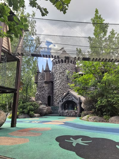 This media file depicts a whimsical, castle-themed playground designed for children. The central subject is a multi-turreted castle structure built with rough, stone-like textures, giving it a fantastical appearance. In front of the castle, a cave-like entrance with a thatched roof is visible, adorned with tree-like carvings. The ground in the foreground is a soft, multi-colored rubberized surface with playful, organic shapes and patterns, suggesting a safe and engaging play area.

Above the castle, a net bridge spans the scene, connecting to wooden structures on either side, hinting at a larger adventure park or complex. Lush green trees and foliage surround the castle and the play area, creating a natural and somewhat enclosed environment. Large, natural-looking boulders are strategically placed around the landscape, adding to the organic and fantastical aesthetic.

While no specific people are clearly visible and engaged in activities, the presence of a playground and the castle structure strongly imply that children are the intended users and likely users of this space. The scene appears to be set during the daytime, with a mix of sunlight and clouds visible in the sky, suggesting fair weather. The overall mood is one of fun, imagination, and adventure, designed to spark a child's fantasy. The location context provided is Sovkhoza Im. Lenina, Russia, which places this playground within that specific geographical area. There is no visible text in the image.