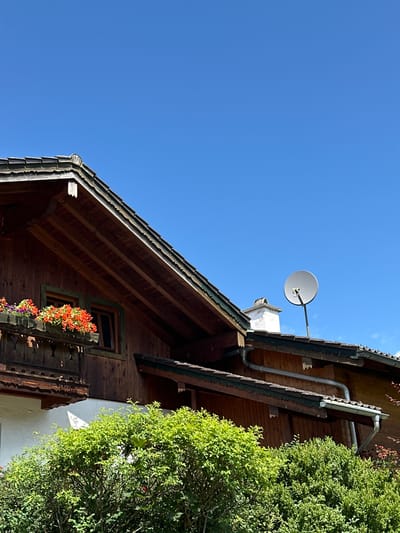An exterior, eye-level, medium shot captures a portion of a wooden, multi-story building against a clear, bright blue sky. The building features a sloping, dark-wood shingled roof with exposed wooden rafters and decorative eaves. A wooden balcony with ornate trim is visible on the left side, adorned with planters filled with vibrant red and purple flowers. Below the balcony, a white wall is partially obscured by lush green bushes. To the right, a satellite dish is mounted on the roof. The scene is set in Bischofswiesen, Germany, suggesting a mountainous or rural environment. No people are present.