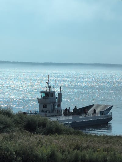 A ferryboat with several people on its deck is seen on the water, with a forested shoreline in the foreground and a hazy horizon in the background. The water is shimmering with sunlight.
