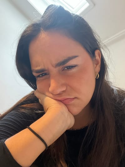 A young adult female is depicted in a close-up indoor shot, looking directly at the camera with an expression of displeasure or weariness. Her dark hair is parted roughly in the center and falls over her shoulders. Her forehead is furrowed, eyebrows are drawn together and down, eyes are narrowed, and the corners of her mouth are downturned. Her left hand supports her chin and jawline. She wears a dark, possibly black, long-sleeved top, a black elastic band on her left wrist, and a small, beaded gold-toned hoop earring on her visible left ear. The background is out of focus, revealing a plain, light-colored wall and a rectangular light source, likely a skylight or window, at the top center. No specific visual cues are present in the image to identify a precise location within the city of Bath, United Kingdom.