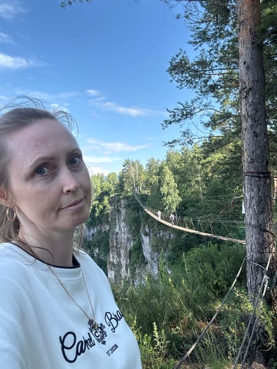 This is a vertical, selfie-style photo taken outdoors. The main subject is a woman with blonde hair and blue eyes, who is looking at the camera. She is wearing a white t-shirt with black lettering and a gold necklace. The background shows a lush green forest, a rocky cliff face, and a suspension bridge spanning across a gorge. The sky is bright blue with a few wispy clouds. The overall impression is that of a sunny day, possibly late morning or early afternoon. The woman's expression appears relaxed and perhaps a little adventurous, suggested by the dramatic natural landscape behind her.
On the woman's t-shirt, there is visible text that appears to read "Cali Bro" and "ST. 2025" with a decorative emblem. The scene suggests an outdoor recreational activity, possibly hiking or exploring, with the suspension bridge indicating a location that offers such activities. The natural elements like the large tree trunk in the foreground and the dense foliage imply a natural park or wilderness area. The image is taken in portrait orientation and appears to be a personal snapshot.