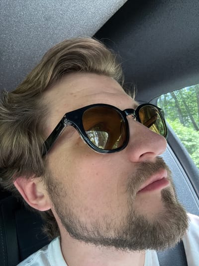 The image shows a close-up, side profile of a person wearing sunglasses and a white t-shirt, likely inside a vehicle. The person has light brown hair, a light beard, and is looking to the right. The sunglasses have black frames and amber lenses, reflecting trees and sky. The interior of the vehicle, including the headliner and part of a seat, is visible. The scene is outdoors, with greenery and trees visible through the vehicle window. The location is identified as Krubki-Górki, Poland.