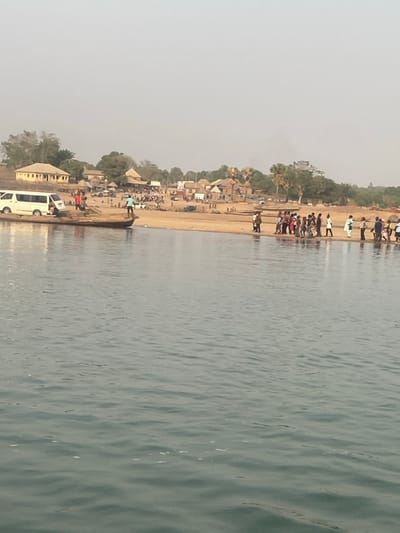 A landscape captures a shoreline with a sandy beach lined with various structures and trees. People are visible along the beach. The beach is bordered by a body of water. A white van is parked near a building at the lower edge. The sky appears hazy.