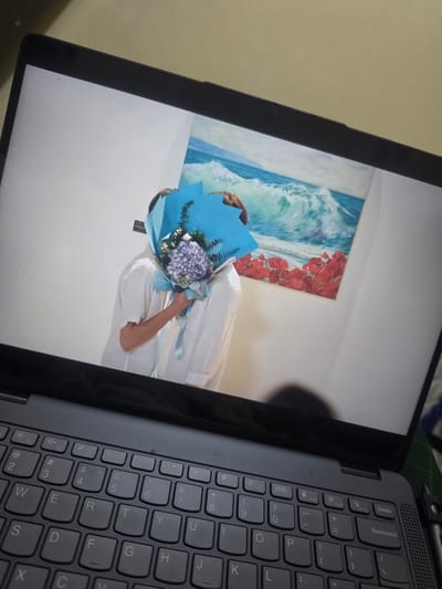 The image depicts a laptop screen displaying a photograph. The photograph shows a person holding a bouquet of flowers wrapped in blue paper. In the background of the photograph is a wall with a painting of ocean waves and red flowers. The laptop's keyboard is visible in the foreground. The scene appears to be indoors. The image was taken in Pintomakmur, Indonesia.