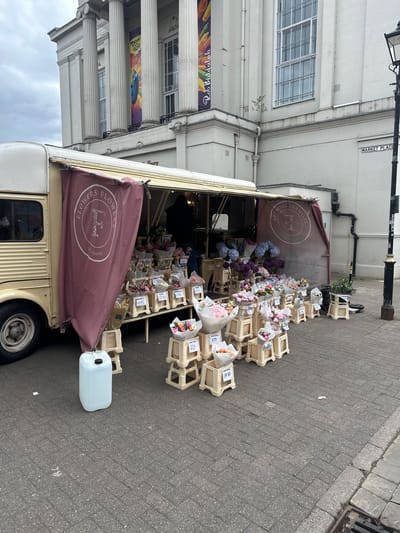 The image shows a flower stall situated next to a building with classical architectural elements, including columns and large windows. A sign reading "MARKET PLACE" is visible on the building. The stall is a van-like structure with a canopy displaying the text "FLOWERS FLOWERS ST. ALBANS" in a circular logo. Numerous bouquets of flowers are arranged in wooden crates, with price tags displayed on some of them. A white plastic container is positioned beside the stall. The scene is shot at street level on a paved area.