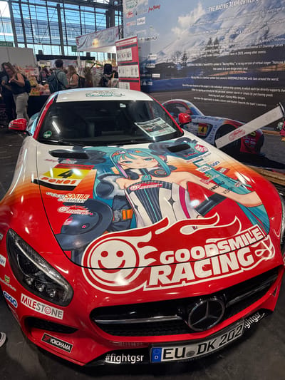 The image shows a red Mercedes-Benz car with a large anime-style graphic on its hood, parked indoors. The car is heavily branded with "GOODSMILE RACING" text and logos. In the background, there are booths and displays associated with a convention or exhibition, suggesting a public event. People are visible in the background, browsing the exhibits. The context provided indicates this scene is from the Paris Manga & Sci-Fi Show at the Paris-Nord Villepinte Exhibition Centre in Villepinte, France. The car itself is the primary subject, adorned with vibrant graphics of an anime character, likely Hatsune Miku, a virtual singer often associated with racing teams. The car's headlights, grille, and license plate are clearly visible. The environment appears to be an exhibition hall with bright lighting and a diverse crowd.