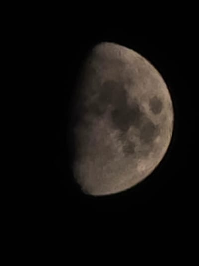 The image shows a close-up of the Moon against a dark, black sky. The Moon is partially illuminated, displaying distinct craters and surface features. The light source is to the right of the frame, casting a shadow on the left side of the Moon, indicating a crescent or gibbous phase. The image is grainy and appears to be taken at night, as suggested by the dark background. There are no people, objects, or discernible location cues in the image other than the celestial body itself. The setting is outer space, specifically focusing on the Moon. No text is visible in the image.