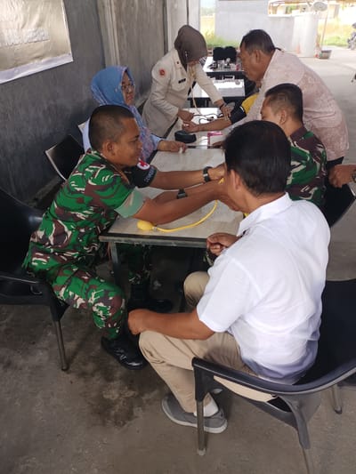 In a shaded outdoor area with a concrete floor, a group of individuals appears to be engaged in a medical check-up, possibly a blood pressure measurement, in Cotgadong, Indonesia. In the foreground, a person in a white short-sleeved collared shirt and khaki pants sits in a black plastic chair, facing away from the camera. Their left arm is extended, and a blood pressure cuff is being applied by a person in a camouflage military uniform. The soldier, also seated in a black plastic chair, wears a green, brown, and black camouflage uniform and black boots. A yellow bulb and tubing, likely part of a sphygmomanometer, are visible.
Behind these two individuals, a person in a beige hijab and a light-colored uniform with a red cross emblem is seated, observing the procedure. To their right, another person in a light-colored patterned shirt stands, leaning over the table. A woman in a blue hijab and glasses sits on the left, looking towards the activity. The scene suggests a community health initiative or a military medical service being provided in an open but covered space, indicated by the concrete floor and the presence of a building structure behind the participants. A banner with text, partially visible on the left, is attached to the wall. The overall atmosphere appears calm and procedural.