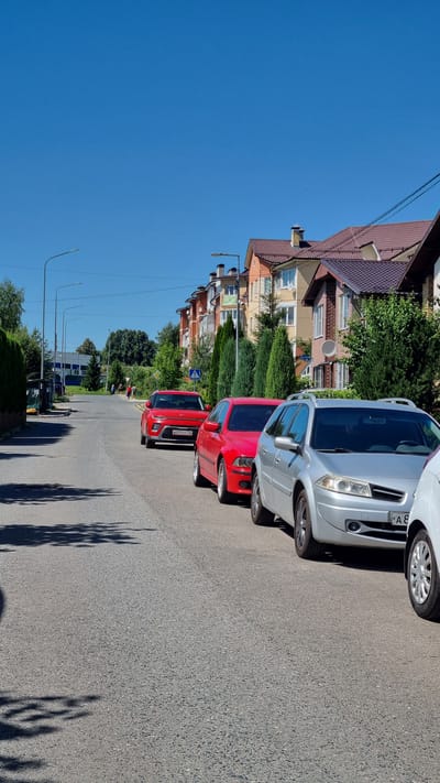 The image depicts a street scene in the city of Verzilovo, Russia, on a clear, sunny day. In the foreground, several cars are parked along the side of an asphalt road. The closest car is a silver station wagon, followed by a red sedan and a red SUV. The buildings lining the street are modern, multi-story residential structures with pitched roofs. Several evergreen trees are planted along the sidewalk in front of the buildings. In the background, the road continues, with more buildings and trees visible. There are a few pedestrians walking on the sidewalk in the distance. The sky is a vibrant blue.