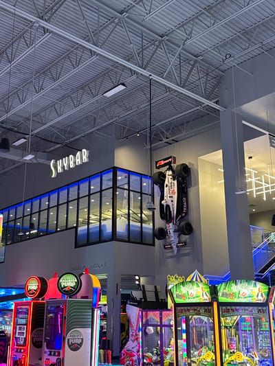 This vertical, wide-angle shot captures a lively indoor entertainment complex in Schaumburg, United States of America. The high ceiling features an industrial design with exposed white metal trusses and corrugated panels.
A striking white race car, appearing to be a replica of an IndyCar or Formula 1 vehicle, is suspended from the ceiling. It prominently displays sponsor logos such as "TEXACO," "smart," "Energizer," and "Gillette," along with the number "1" and "GOODYEAR" on its tires. Other visible text on the car includes "CORNING" and "Pit Devil BASE."
To the left, a large, illuminated sign reads "SKYBAR," indicating a bar area. Below this, a modern architectural feature with square glass panes is highlighted by blue neon-like lighting. Further right, a sign for "VKARTING TRACKY" (likely "KARTING TRACK") suggests a go-karting facility.
The right side of the image is dominated by a vibrant array of arcade games. Several machines feature "PERFECT PUMP" branding with circular, illuminated signs. Another prominent game is "The WIZARD Emerald City Edition," with visible text encouraging players to "Collect all cards to receive a point bonus!" Other faint text seen includes "NFS," "LAIGAMES," and "NASDAQ." The bright lights and varied attractions indicate a bustling recreational environment. The indoor setting means the time of day is not discernible.