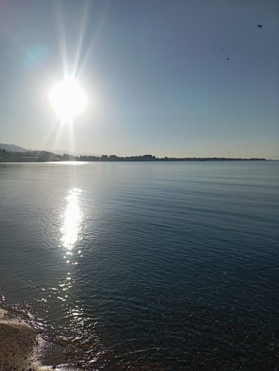 The image depicts a serene lake scene at sunrise. The sun, a brilliant orb with distinct rays, shines brightly in the upper left quadrant of the sky, casting a shimmering reflection across the calm water. In the distance, a faint outline of hills and a treeline with buildings suggest a populated area. The water itself is a deep blue, with subtle ripples creating a textured surface, and the shallow area near the shore reveals a bed of pebbles. A few small birds are visible in the clear blue sky. The overall impression is one of natural beauty and tranquility. This scene is consistent with a view of Lake Issyk-Kul from the shore in Bosteri, Kyrgyzstan, a popular resort area known for its clear waters and mountain backdrop.