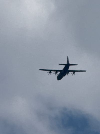 A silhouetted cargo airplane, likely a Lockheed C-130 Hercules, flies against a cloudy sky in Dolni Bogrov, Bulgaria. The aircraft is dark and appears to be in flight, with its four engines visible under its wings. The sky is overcast, with varying shades of grey and white clouds. The image is a low-angle shot, looking up at the plane from below.