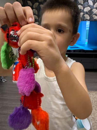 The image shows a close-up of a young boy holding a string of colorful, fluffy pom-pom charms. He is wearing a white t-shirt and his hands are in focus as he manipulates the charms. The boy's face is partially visible, with his eye looking down at the objects. The background is blurred but suggests an indoor setting, possibly a home, with a patterned curtain on the left and a television screen in the upper left corner. The lighting appears to be natural and warm, indicating it might be daytime. The scene conveys a sense of focus and play as the boy interacts with the charms. No visible text is apparent on the charms or in the background. The provided location context is Malmyzh, Russia.
