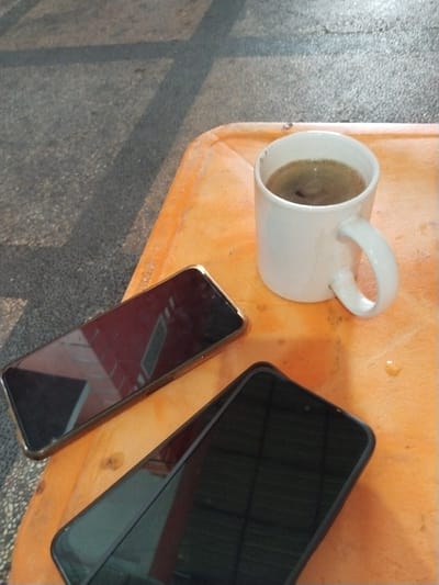 The image depicts two black smartphones and a white mug of coffee placed on an orange plastic tray. The tray rests on a speckled gray surface, possibly a sidewalk or pathway, with shadows cast across it, indicating an outdoor setting. The scene is likely situated in an outdoor cafe or public space in Pekanbaru, Indonesia, given the typical weather and urban environment found there. The smartphones are positioned flat on the tray, with their screens reflecting the surroundings. The coffee mug is also on the tray, filled with dark coffee. The overall impression is that of a casual break or moment of relaxation.