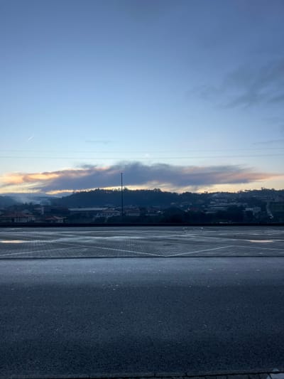 A striking view showcases a blend of natural and built environments. On the left, a blue sky hosts elongated, textured clouds that transition from light to dark tones. A distant line indicates an airplane trail. The skyline features structures nestled against the edge of a dark hillside. The right side features an area covered in dark material, possibly asphalt or a similar paving surface. The scene evokes a quiet, possibly dawn or dusk, atmosphere.