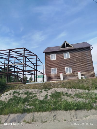 An exterior, eye-level, wide shot captures a construction site next to a wooden house under a blue sky with scattered clouds. On the left, a large, unfinished steel frame structure, likely for a building, dominates the foreground. To the right, a two-story, dark brown wooden house with a brown tiled roof and a triangular gable window is visible. The house has white-framed windows with metal grates on the lower level. A light-colored metal fence with white posts runs in front of the house. The ground in the foreground consists of a dirt embankment with sparse green vegetation. A white sign with green lettering is partially visible behind the steel structure. The timestamp "07.07.2026 16:43" and "REDMI Note 13" are superimposed on the bottom of the image. The scene suggests ongoing construction and development in a semi-rural or suburban area.