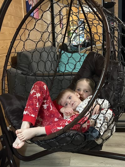 Two children are seated in a hanging pod chair indoors. One child is wearing red pajamas with white Christmas trees and reindeer, while the other child is wearing pajamas with a milk bottle print. The chair is dark brown with a mesh structure and has a dark brown cushion. The room has a wooden floor. A couch with throw pillows can be partially seen in the background. There is no indication of the location being Bol'shakino, Russia.