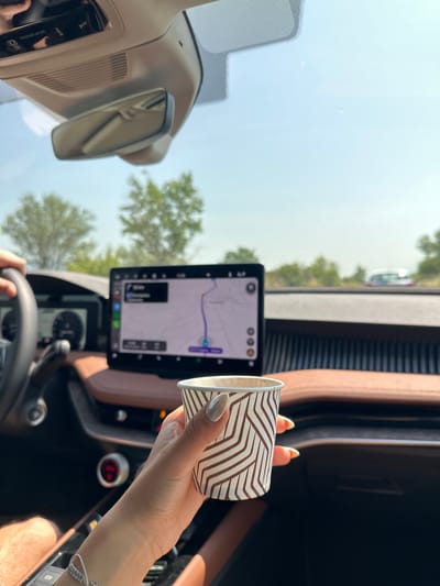 The image shows the interior of a car, possibly a modern luxury vehicle given the large touchscreen display and brown leather accents. A person's hand, with painted fingernails, is holding a disposable coffee cup with a brown and white patterned design. The car's dashboard is visible, including the steering wheel, a digital instrument cluster, and the central infotainment screen displaying a navigation map. Outside the car, a bright, sunny day is evident with trees and a blue sky. There are hints of other vehicles on the road in the distance. The scene appears to be set on a highway or main road, given the context of navigation and the presence of other cars. There is no explicit indication of the location being Chryso, Greece, but the visual elements are consistent with a roadside scene in a temperate climate.