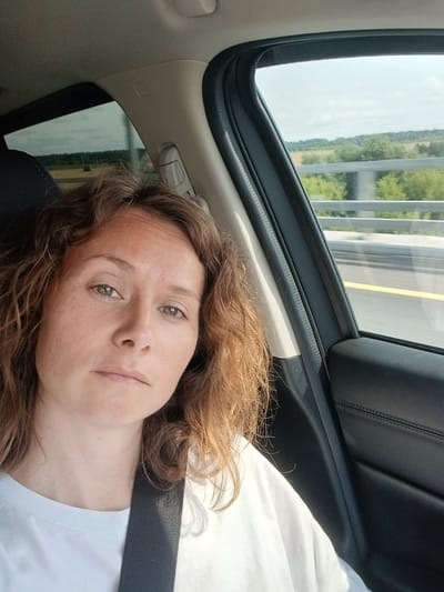 In Pilekshevo, Russia, a woman with shoulder-length wavy brown hair is photographed from the chest up, facing the camera while seated in a car. Her gaze is directed forward, with a neutral expression. She is wearing a white t-shirt and a dark seatbelt is visible across her chest. The interior of the car, including a black leather door panel and a beige headliner, is visible. Sunlight streams through the car window, illuminating the scene. Outside the window, a blurred landscape of green trees and fields, along with a highway railing, suggests the car is in motion. The sky is overcast with scattered clouds.