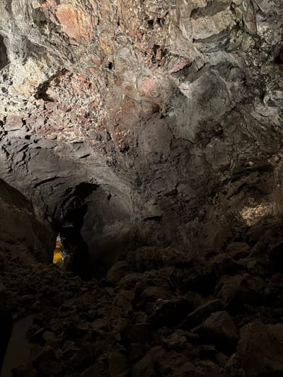 The image depicts the interior of the Cueva de los Verdes, a lava tube located in Haría, Lanzarote, Spain. The scene is largely dominated by the cave's rocky, textured walls, illuminated unevenly by artificial lighting, creating areas of bright exposure and deep shadow. The rock varies in color from reddish-brown to dark gray. In the distance, a small opening glows with artificial light. No people or other specific subjects are visible in the frame.