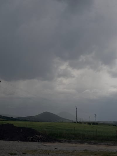 Overcast sky dominates the landscape in Begunovtsi, Bulgaria. Power lines run through the scene, supported by utility poles. A green field stretches towards a distant hill, adjacent to a rocky roadside.
