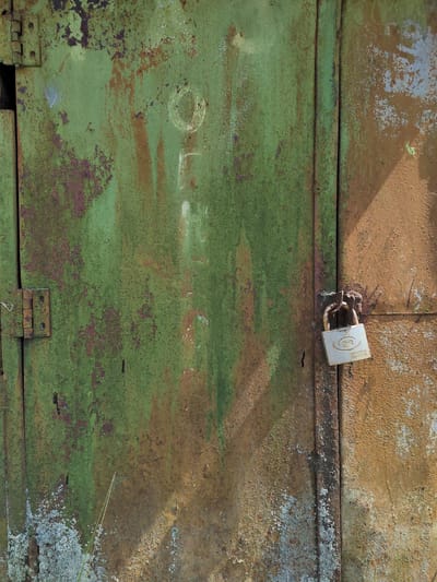 A close-up, full-frame, outdoor vertical shot displays a weathered, green, and rust-colored metal door with a locked padlock. The door appears to be part of a garage or storage unit. The green paint is peeling in numerous places, revealing patches of rust and an underlying material. The right side of the door is primarily a rusty orange-brown hue with lighter, flaky areas. A metal hinge is visible on the left side of the door. A silver padlock is attached to a metal loop, securing the two vertical panels of the door. The padlock has a logo that includes stylized letters and the word "NORAM". Sunlight casts a shadow diagonally across the right side of the door. The environment appears to be within the Бярэзінскі сельскі Савет, Belarus. There are no people or animals visible in the image.