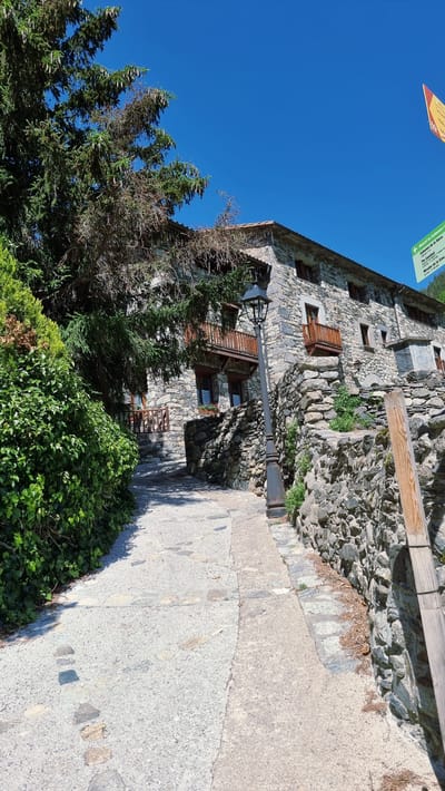 This media file captures a scene in Setcases, Spain, under a clear blue sky, suggesting a sunny day. The main subject is a stone building, characteristic of traditional Pyrenean architecture, with wooden balconies and windows. The building is nestled against a rocky hillside, and a paved pathway, bordered by greenery, leads alongside it.

In the foreground, lush green bushes and trees, including a large evergreen, add to the natural setting. A vintage-style street lamp stands tall beside the building, casting a shadow on the pathway. A wooden beam runs horizontally above the stone wall, possibly part of a fence or a structural element. The pathway itself is made of concrete and cobblestones, showing signs of age and wear.

There are no people visible in the image, and no specific activity or event is taking place. The focus is on the architectural charm and the serene outdoor environment.

At the top left of the image, a partially visible signpost with yellow and green elements is present. Though the text is largely obscured and difficult to read clearly, it appears to be informational, possibly indicating directions or local attractions. The overall mood is tranquil and picturesque, highlighting the rustic beauty of the location.