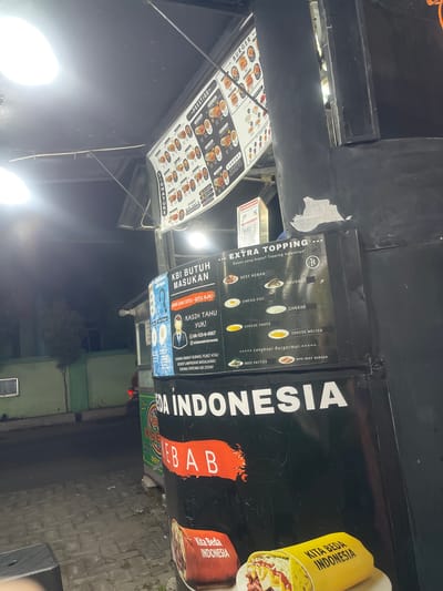 The image shows a food stall at night in Anggaswangi, Indonesia. The stall features a menu board with pictures of burgers and toppings, including beef kebab, sausage, omega egg, cheese, cheese sauce, cheese melted, beef patties, and mini beef burgers. There is also a section for "Extra Topping" and "Signature". A sign above the menu board says "PRESTIGE." Below the menu board, there is a sign that says "KBI BUTUH MASUKAN" which translates to "KBI needs input." The stall is selling burgers and kebabs, as indicated by the text "KITA BEDA INDONESIA" on a kebab wrapper. The stall is brightly lit by artificial lights. There are no people visible in the image, but the scene suggests a commercial establishment. The surrounding environment appears to be a street or sidewalk with some trees and buildings in the background.