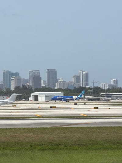 The image depicts an aerial view of an airport in Dania Beach, United States of America, with a city skyline in the background. Two airplanes are visible on the tarmac, one white and one blue with "JetBlue" branding. The airport features multiple runways and taxiways, marked with yellow and white lines. Several airport buildings and structures, including hangars, are also present. The foreground of the image shows a grassy area, likely part of the airport grounds. The sky is clear and pale blue. The overall scene suggests a typical day at a busy airport with ongoing flight operations.