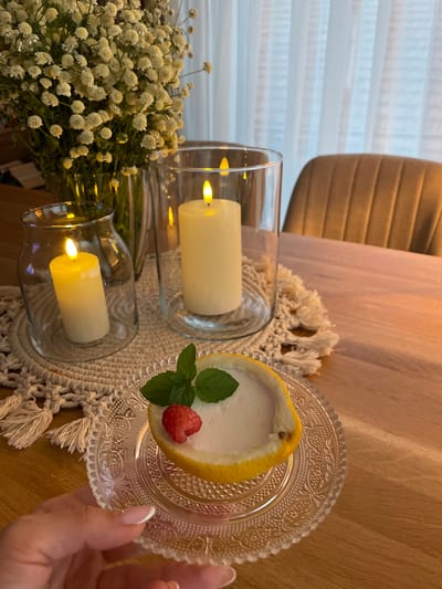 In a dimly lit room in Kukorevo, Bulgaria, a close-up shot captures a wooden table setting. A hand, with manicured nails, holds a small, ornate glass plate. On the plate sits a dessert served in a lemon half, topped with a raspberry and a mint sprig. The dessert itself appears to be a light-colored mousse or cream.

Behind the dessert, two glass candle holders contain flickering LED candles, casting a warm glow. One candle is in a smaller, rounded holder, and the other in a taller, cylindrical one. A macrame placemat with tassels sits beneath the candle holders.

In the background, a bouquet of small, white flowers in a vase adds a touch of natural decor. To the right, the back of a brown upholstered chair is visible, suggesting a dining or living area. White sheer curtains hang in the background, filtering light from outside. The overall atmosphere is cozy and intimate, with the warm candlelight and the dessert creating a sense of indulgence.