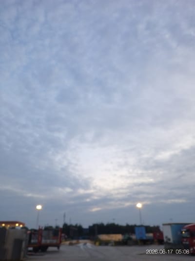 This is a vertical, slightly blurred, outdoor photograph taken at dusk, showing a semi-industrial area. The sky is filled with scattered clouds, predominantly in shades of light grey and pale blue, with a brighter area towards the center indicating the setting sun or ambient light.

In the foreground, a paved area or roadway is visible. To the left, a portion of a light-colored, industrial-style building or structure is partially cropped. Further to the right, a red flatbed truck is parked, with its trailer carrying what appears to be large, grey rectangular objects. Another truck, possibly a semi-trailer with a blue container, is visible behind it, and further to the right, the front of a red truck cab is present. In the mid-ground, a tall lamppost with a glowing light is positioned to the left of center. Behind these vehicles, a line of trees forms a dark silhouette against the sky. The overall environment suggests a logistics yard, a construction site, or an industrial park.

The timestamp in the lower right corner reads "2026.06.17 05:08", indicating the date and time the photograph was taken. The location is Houthalen, Belgium. There are no people visible in the image.