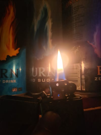 A close-up, low-angle shot depicts a lit Zippo-style lighter in the foreground. The lighter's flame emits a bright yellow and blue light. Behind the lighter, out-of-focus, are two cans of "BURN" energy drink. The can on the left has a blue and teal design with text reading "RN," "DRINK," and "449 ML." The can on the right is darker with text that is partially obscured but appears to say "BURN" and "ZERO SUGAR." In the background, a blurry image of flames or colorful smoke can be seen. The overall lighting is dim, with the flame of the lighter being the primary light source. There is no indication of the city of Gukovo, Russia, in the image.