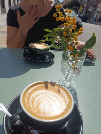A woman is sitting at an outdoor table at a cafe in El Port De Sagunt, Spain. There are two cups of coffee on the table, each in a black cup and saucer. A small, clear glass vase filled with water and sprigs of yellow mimosa flowers sits between the coffee cups. The woman is wearing a black shirt. A car is parked on the street in the background. Buildings line the street.