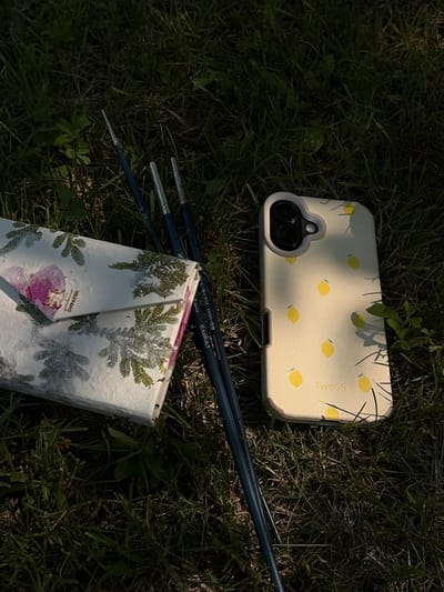 A close-up, high-angle shot shows art supplies and a phone case arranged on a grassy surface. In the foreground, to the left, lies a rectangular object wrapped in white paper adorned with green fern leaves and a pink heart-shaped embellishment. To the right of the paper object, three paintbrushes with dark blue handles are laid diagonally, their bristles pointing towards the top right. A light yellow phone case with a pattern of small yellow lemons and the word "TWOSS" printed on it is positioned in the middle right of the frame. The scene is set outdoors, with dappled sunlight illuminating the objects and casting shadows on the grass. The location is not discernible from the image, and there are no people present.